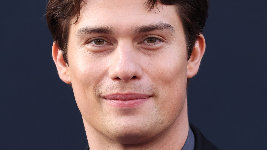 Close-up of actor Nicholas Galitzine smiling and looking directly at the camera, showing his brown eyes, dark hair, and light stubble.