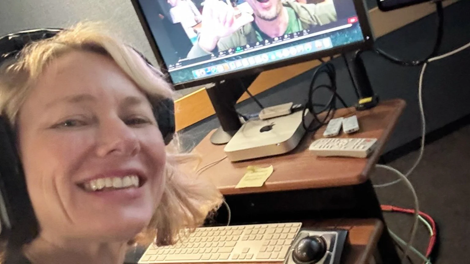 Naomi Watts, wearing headphones, smiles at the camera in a studio setting, with a computer monitor displaying a person and a Mac mini, keyboard, and trackball on a wooden desk.