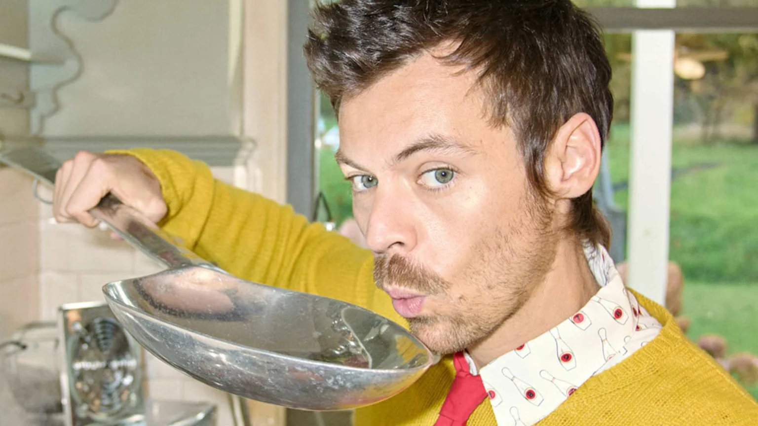 Harry Styles, wearing a yellow sweater and a patterned shirt, holds a large silver spoon to his puckered lips while looking at the camera.
