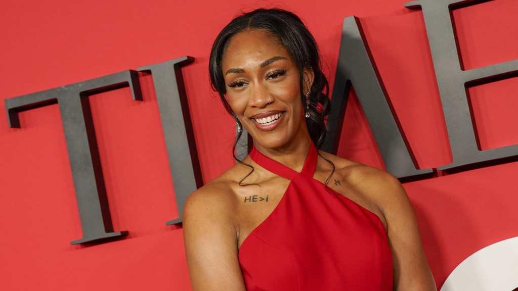 A smiling woman with dark hair, wearing a red halter-neck dress and a "HE>i" tattoo, stands in front of a red background with large "TIME" letters.