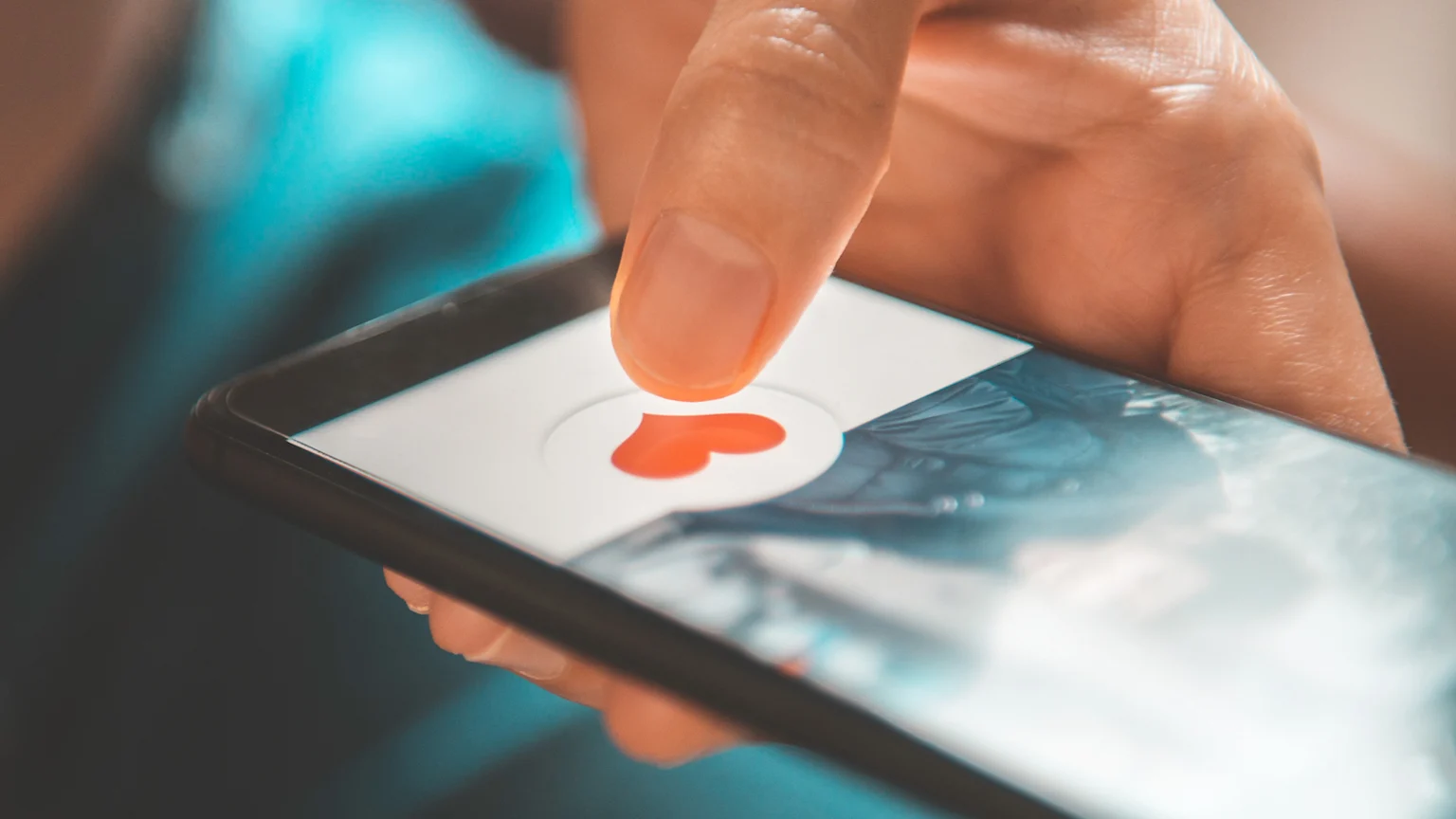 A close-up indoor image showing an adult index finger tapping a red heart icon inside a white circle on a smartphone screen with a black frame. The scene is softly lit with a blurred blue-toned background in a casual setting.