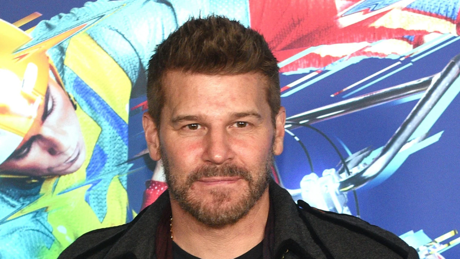 David Boreanaz, with a beard and brown hair, looks directly at the camera while wearing a dark jacket, against a colorful, abstract background.