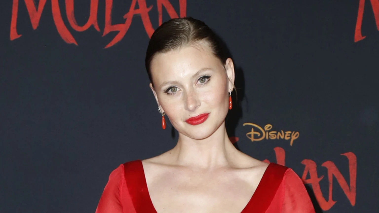 A woman with slicked-back brown hair and red lipstick wears a red dress and red dangle earrings, smiling slightly against a dark background with "Disney Mulan" text.
