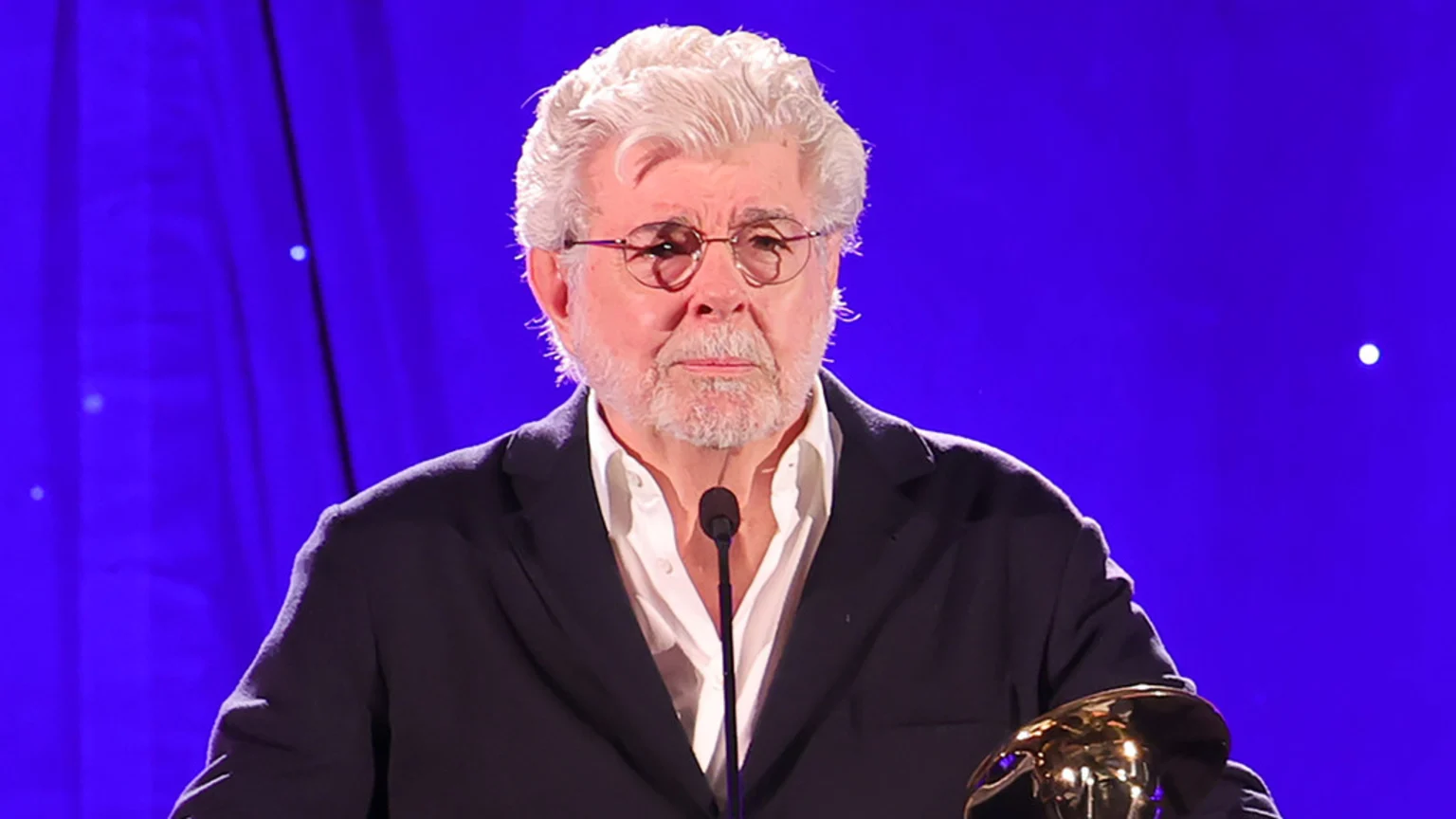 George Lucas, an older man with white hair, beard, and glasses, stands at a microphone wearing a dark jacket and white shirt against a blue background, holding a golden item.