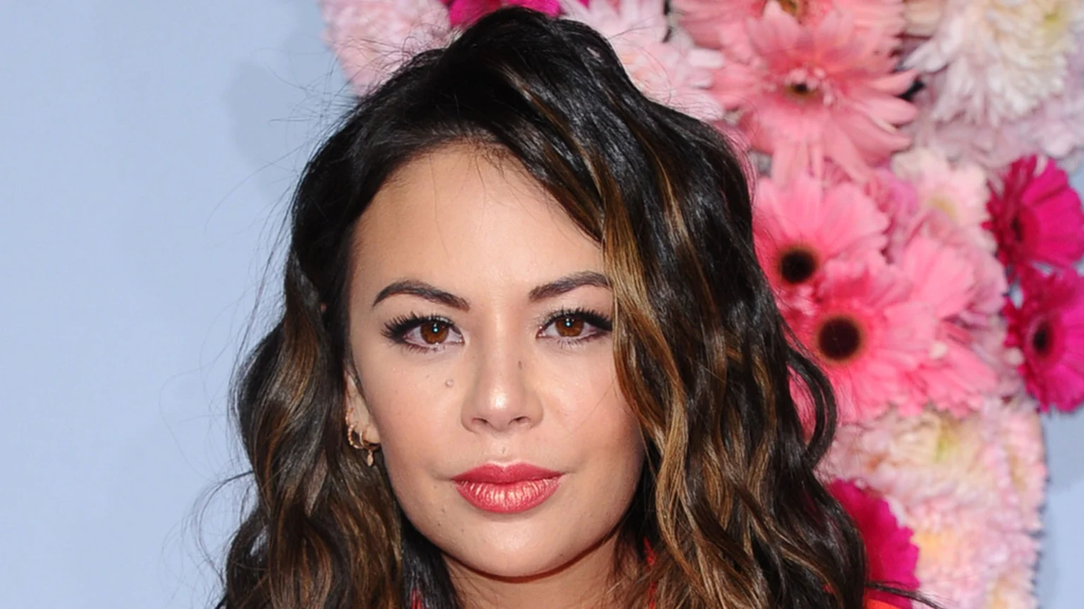 A woman with long, wavy brown hair with highlights, brown eyes, and red lipstick looks directly at the camera, with a background of pink and white flowers.