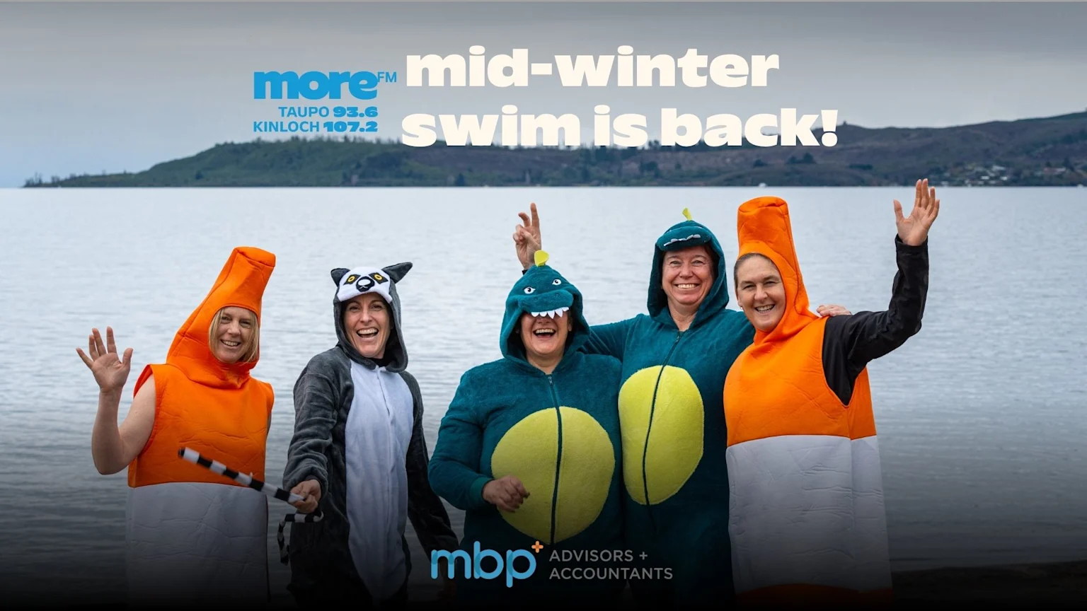 Five people in fun costumes (carrot, lemur, two dinosaurs, traffic cone) wave by a lake, celebrating the "mid-winter swim is back!" event.