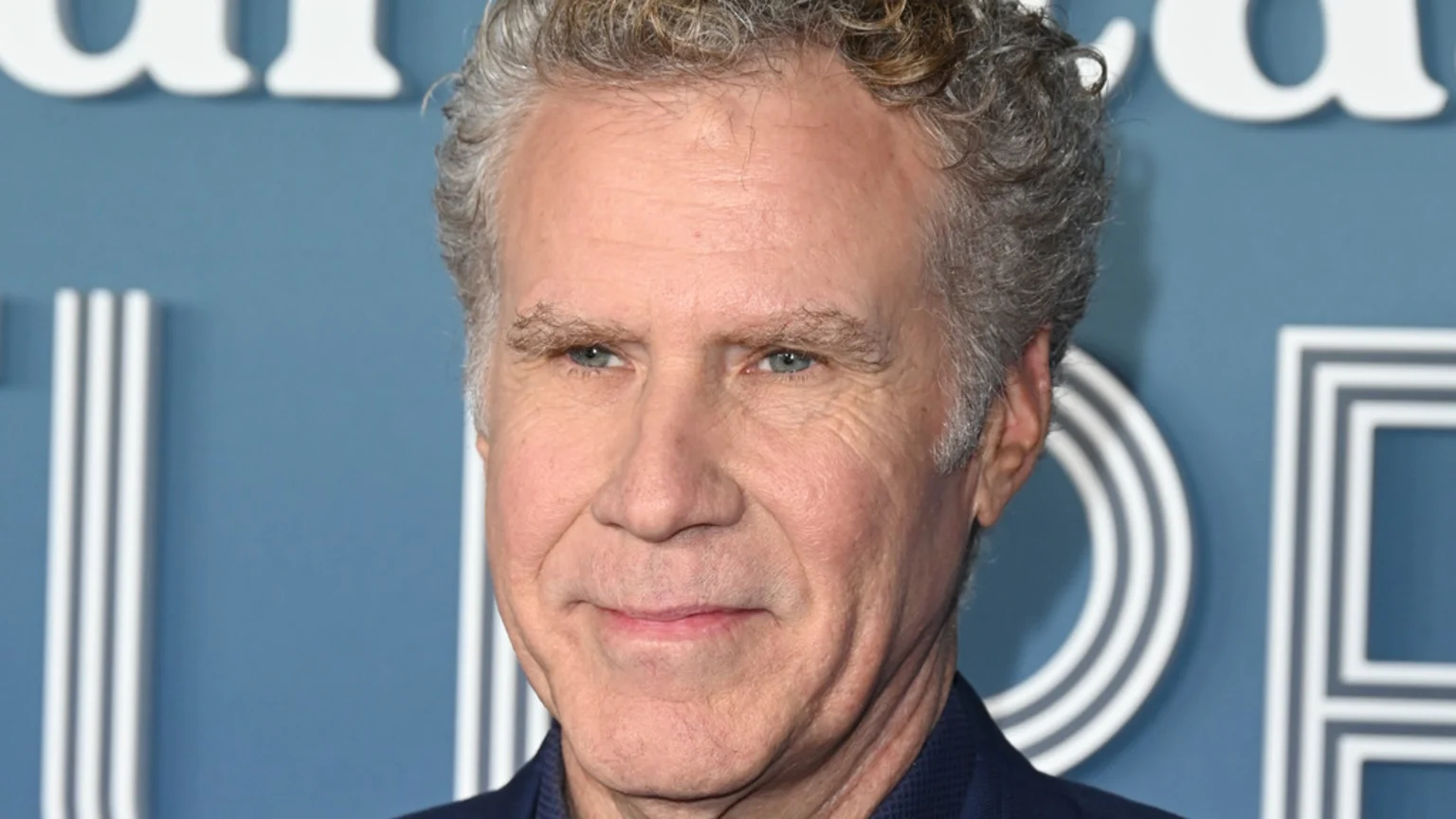 Close-up of actor Will Ferrell with curly grey hair and blue eyes, smiling slightly against a blurred blue background with white architectural details.
