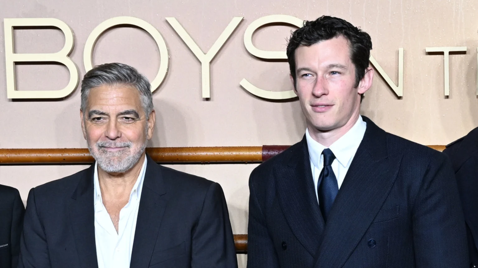 George Clooney, with grey hair and beard, stands smiling in a suit next to Callum Turner, who wears a suit, white shirt, and dark tie, both in front of a light-colored wall with large letters.