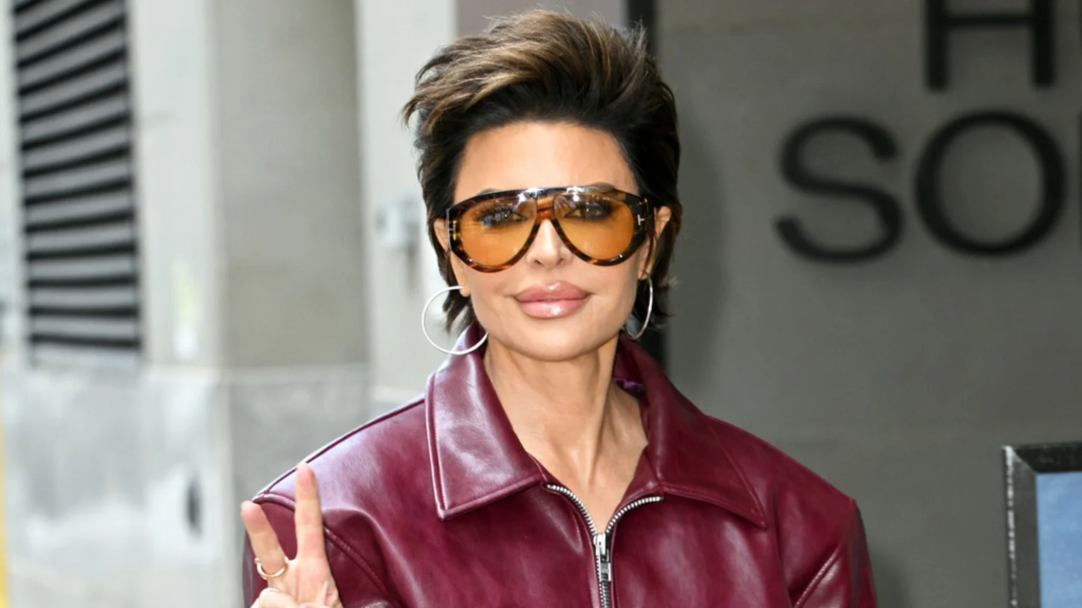 Lisa Rinna with short brown hair, wearing large orange-tinted tortoise-shell sunglasses, a maroon leather jacket, and hoop earrings, makes a peace sign.