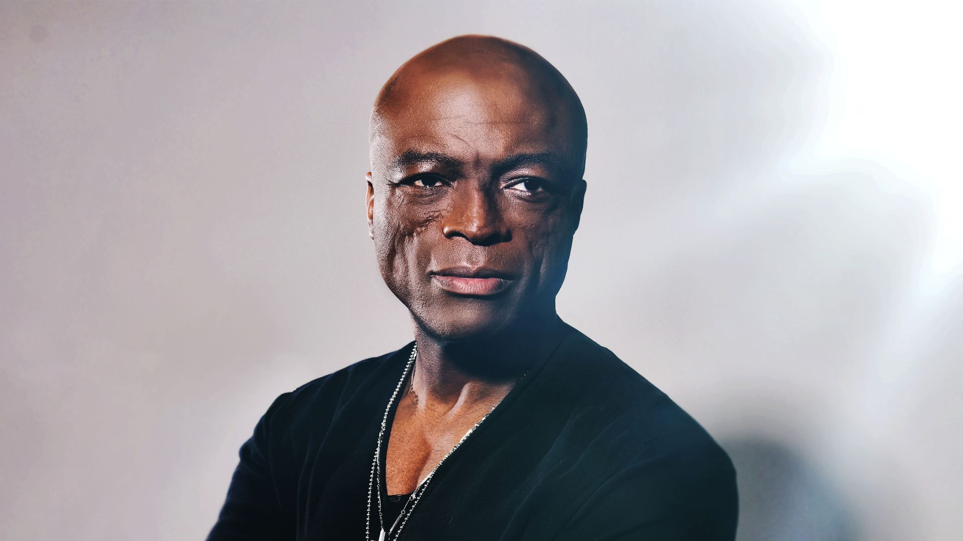 A bald Black man with distinctive facial scars, wearing a black top and a silver chain, looks directly at the camera with a slight smile against a light background.