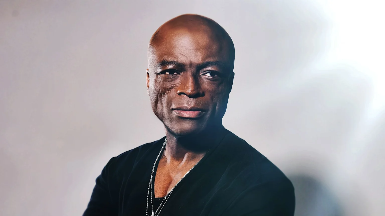 A bald Black man with distinctive facial scars, wearing a black top and a silver chain, looks directly at the camera with a slight smile against a light background.