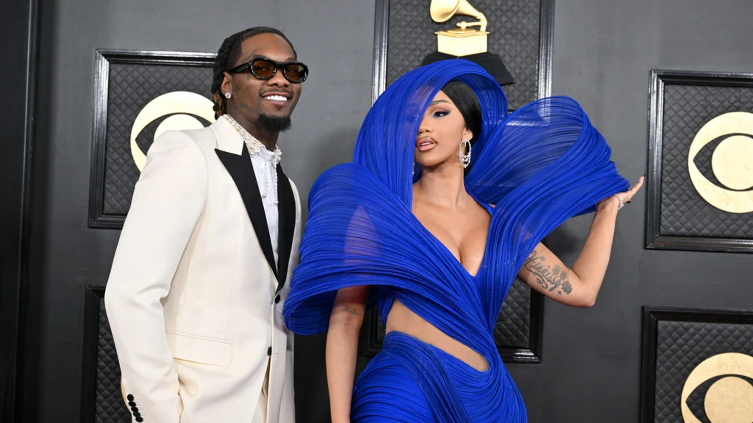 Cardi B in a dramatic blue pleated dress and Offset in a white tuxedo with black lapels smile for a photo in front of Grammy logos.
