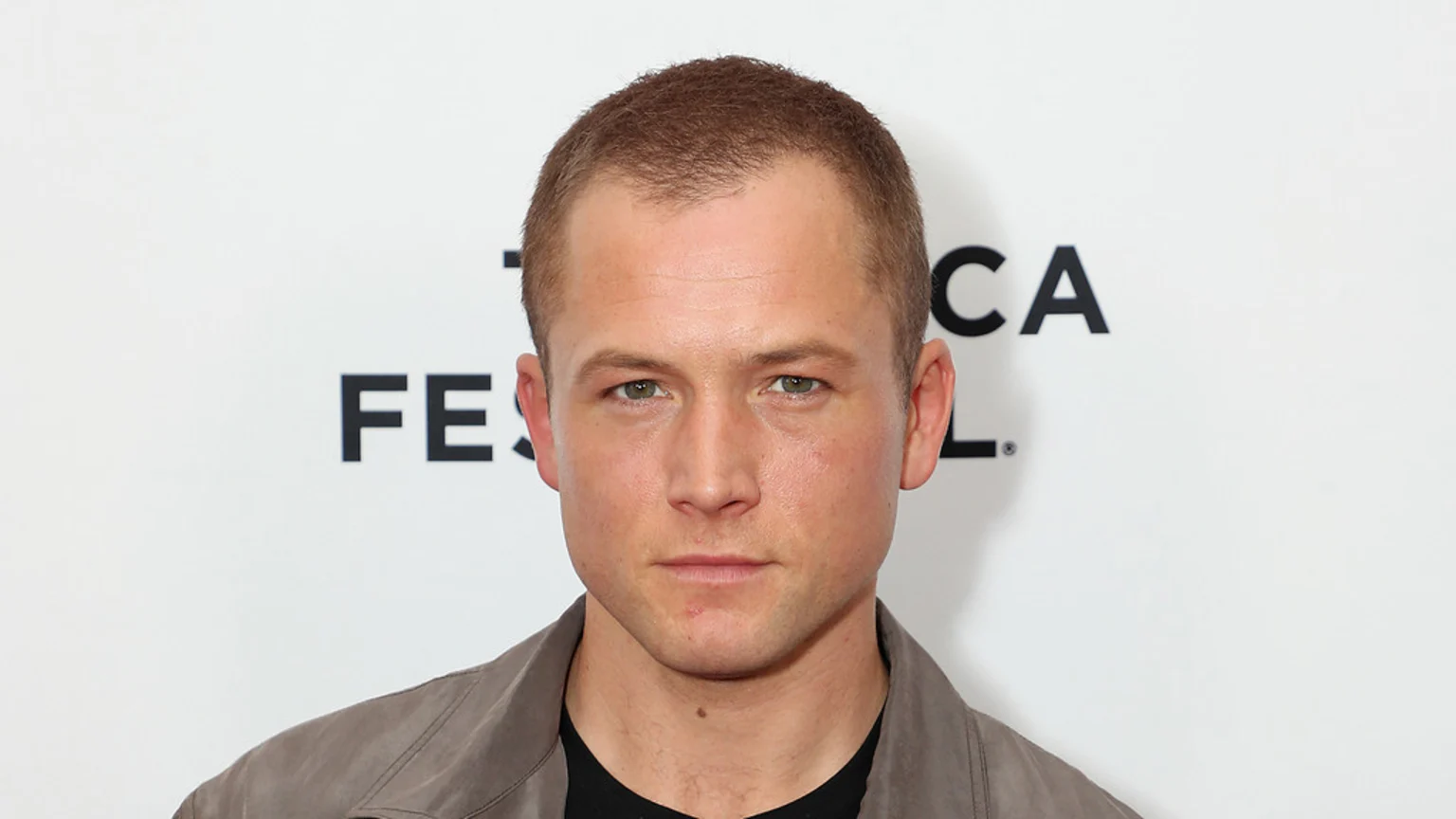 A man with short brown hair and light eyes gazes straight ahead, wearing a dark shirt and light brown jacket, against a white background with partial black text.