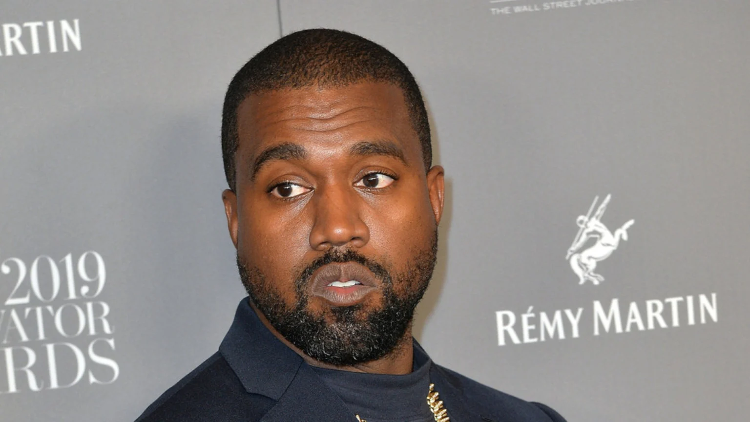 Kanye West, a Black man with a beard, in a dark suit, looks sideways with his mouth slightly open, against a grey background with "2019 INNOVATOR AWARDS" and "RÉMY MARTIN" logos.