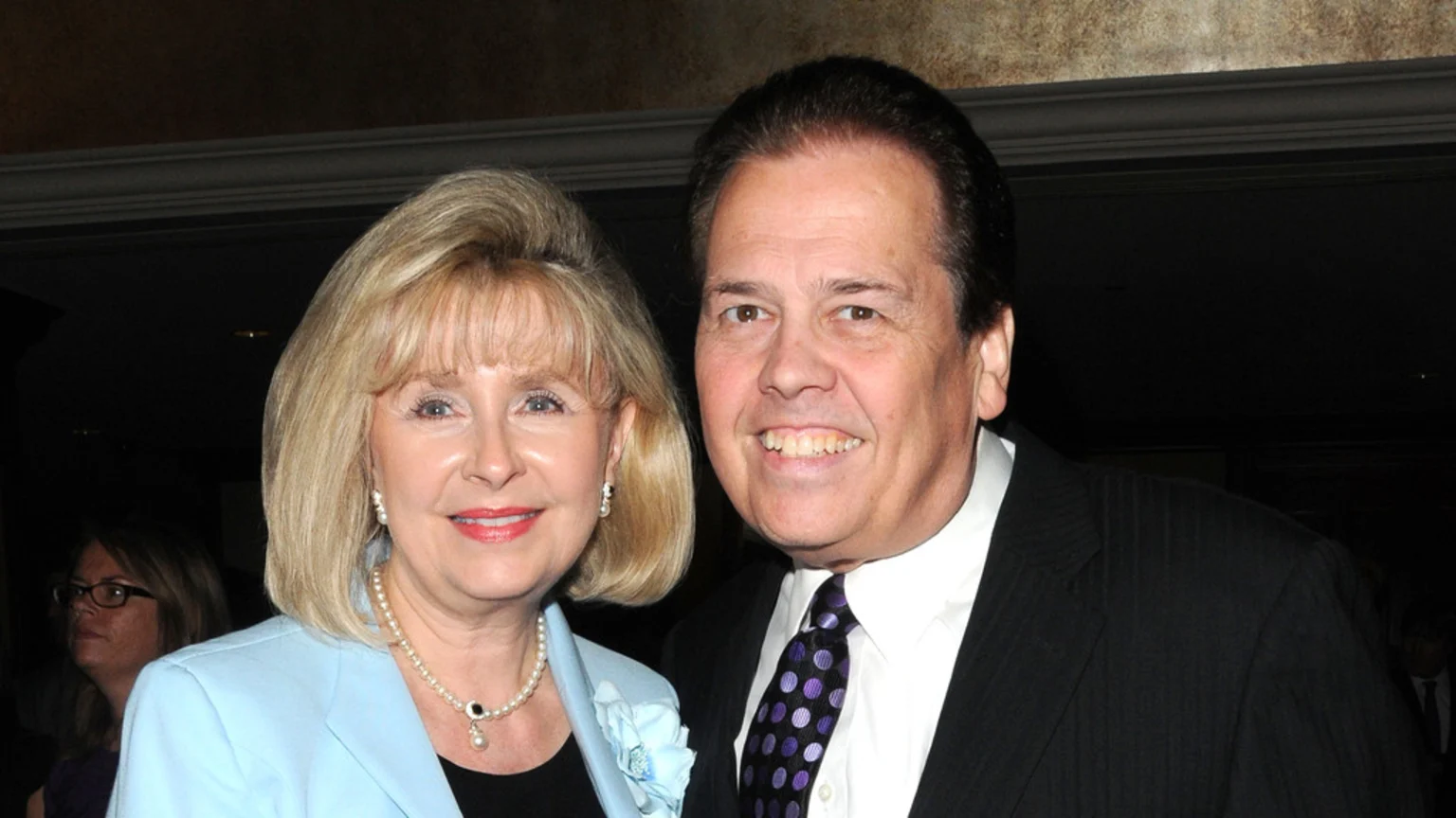 A smiling woman with blonde hair, wearing a light blue jacket and pearls, stands beside a smiling man in a black suit and a purple patterned tie, both looking at the camera.