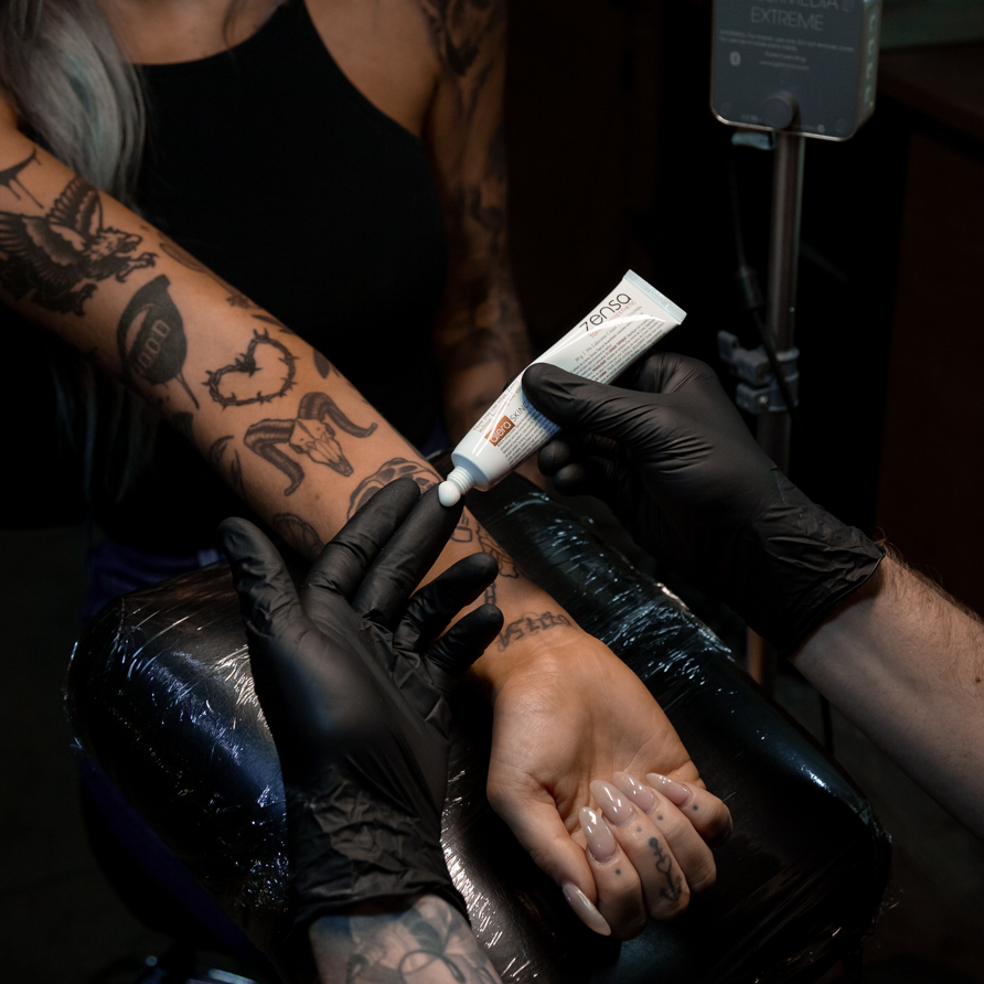 man-apply-zensa-numbing-cream-with-gloved-hands-to-tattooed-girls-arm