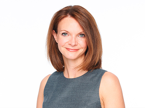 A fair-skinned woman with brown hair, Jeni Thomas, Ph.D., a hair and scalp scientist for more than 20 years, sharing tips on how to reduce hair fall, stands in front of a white background.