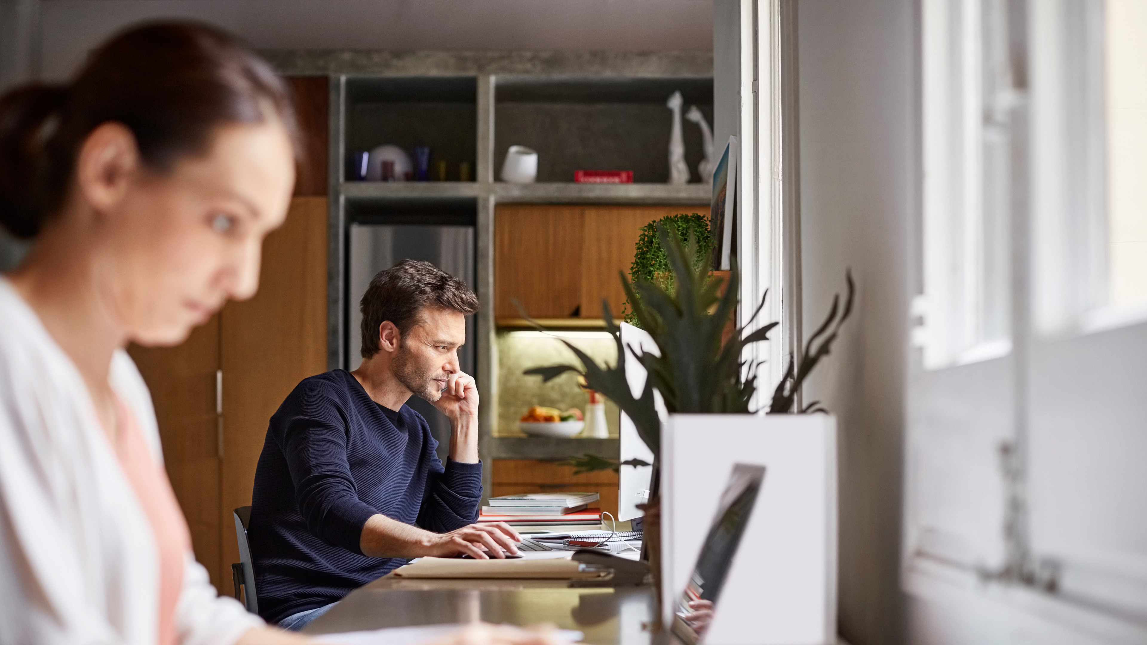 Un ufficio con due persone che lavorano alle scrivanie. Sullo sfondo ci sono scaffali, una pianta e la luce del giorno visibile attraverso le finestre.