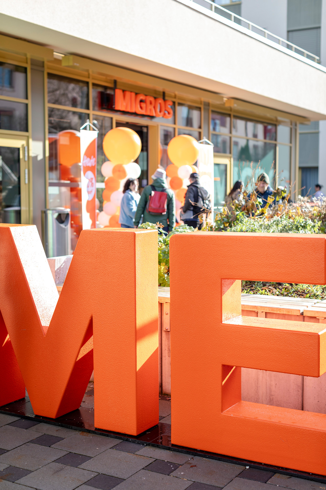 Eingangsbereich einer Migros‑Filiale mit grossen orangefarbenen M‑Buchstaben und festlicher Dekoration vor dem Gebäude.