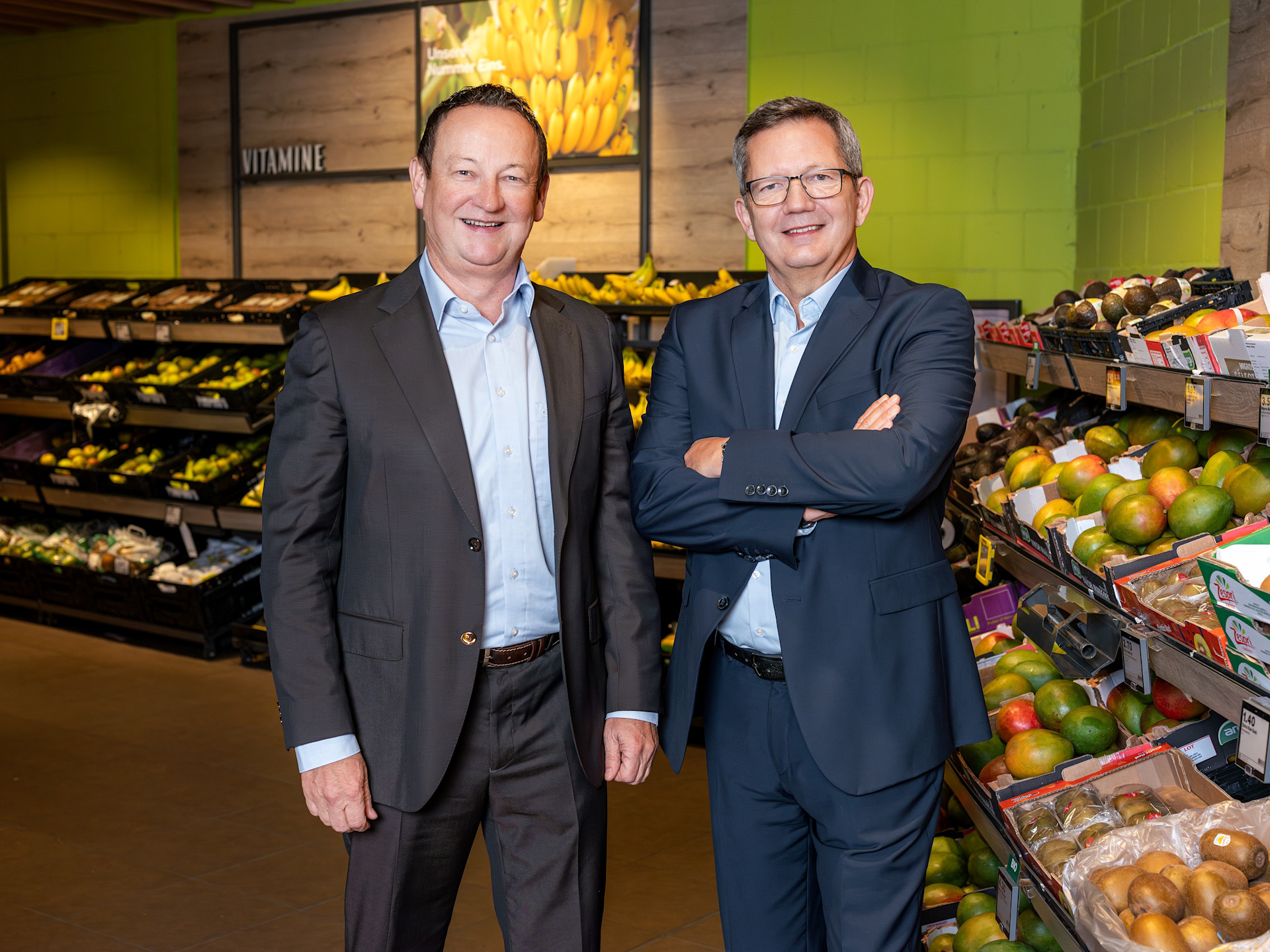 Reto Sopranetti und Jürg Stöckli stehen in Businesskleidung in der Obst- und Gemüseabteilung eines Migros-Supermarkts, umgeben von frischem Sortiment und farbenfrohen Regalen.