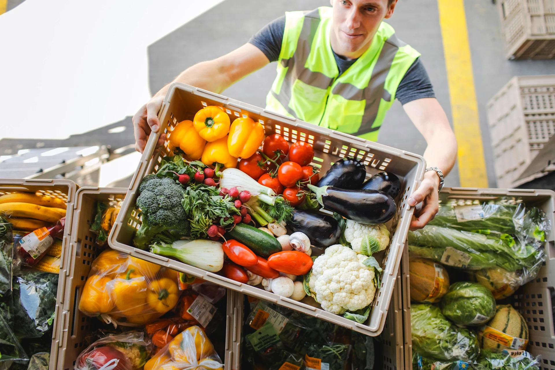 Un homme en gilet de sécurité empile une caisse de légumes multicolores sur d’autres caisses.