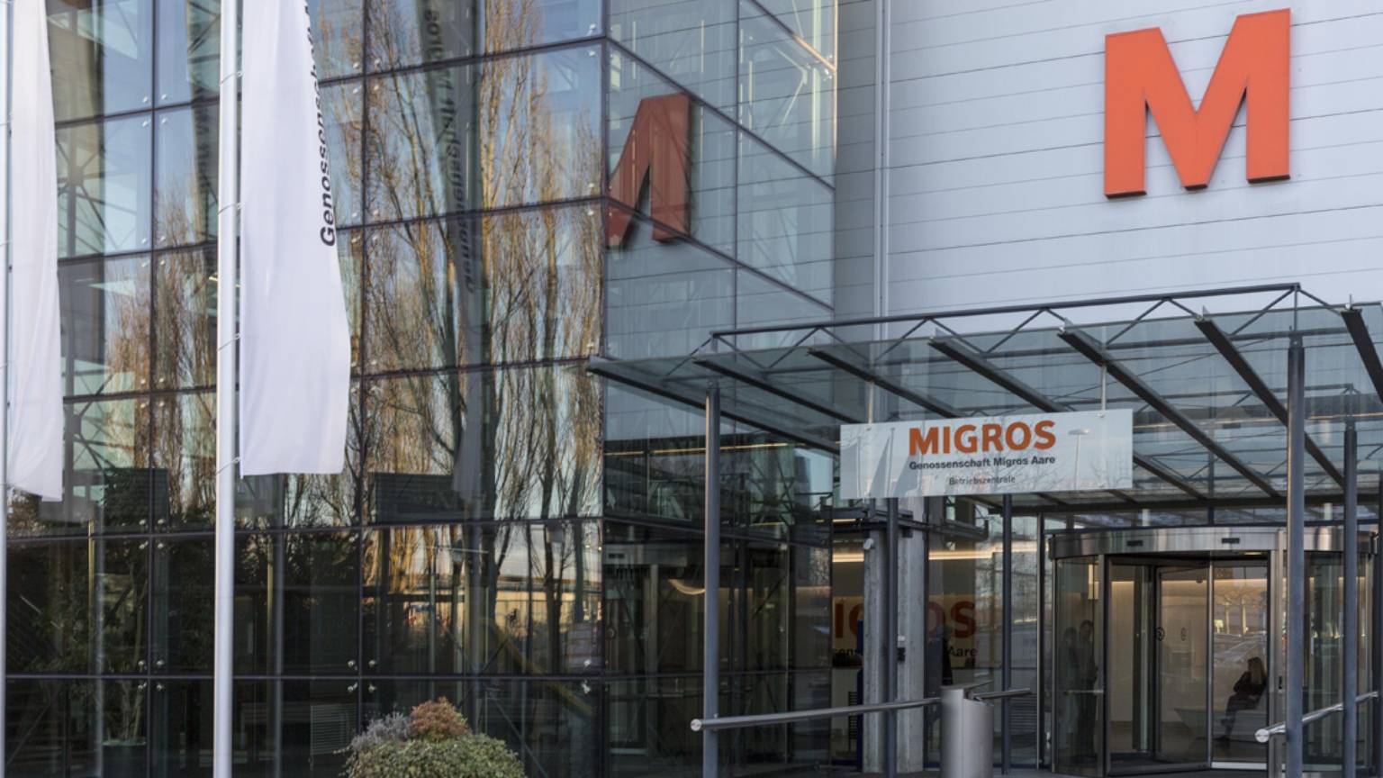 Eingang zum Verwaltungsgebäude der Genossenschaft Migros Aare mit Glasfassade, grossen orangen Migros-Logos und Drehkreuz.