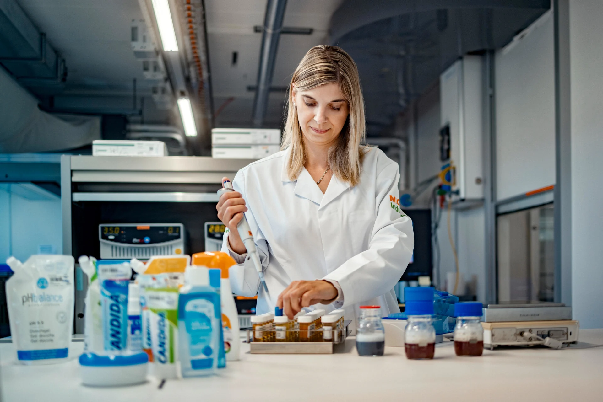 Una donna in camice bianco lavora in piedi in un laboratorio, con vari prodotti Migros davanti a sé.