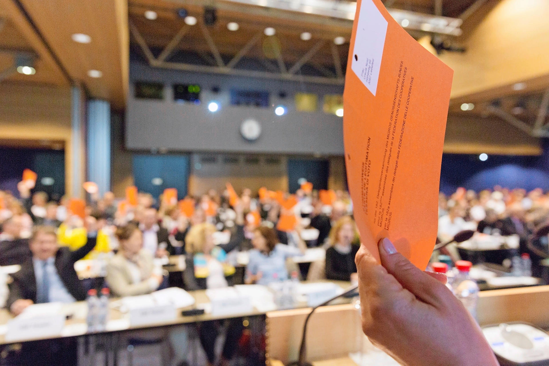In primo piano una mano con scheda di voto arancio, sullo sfondo i membri dell'Assemblea dei delegati.