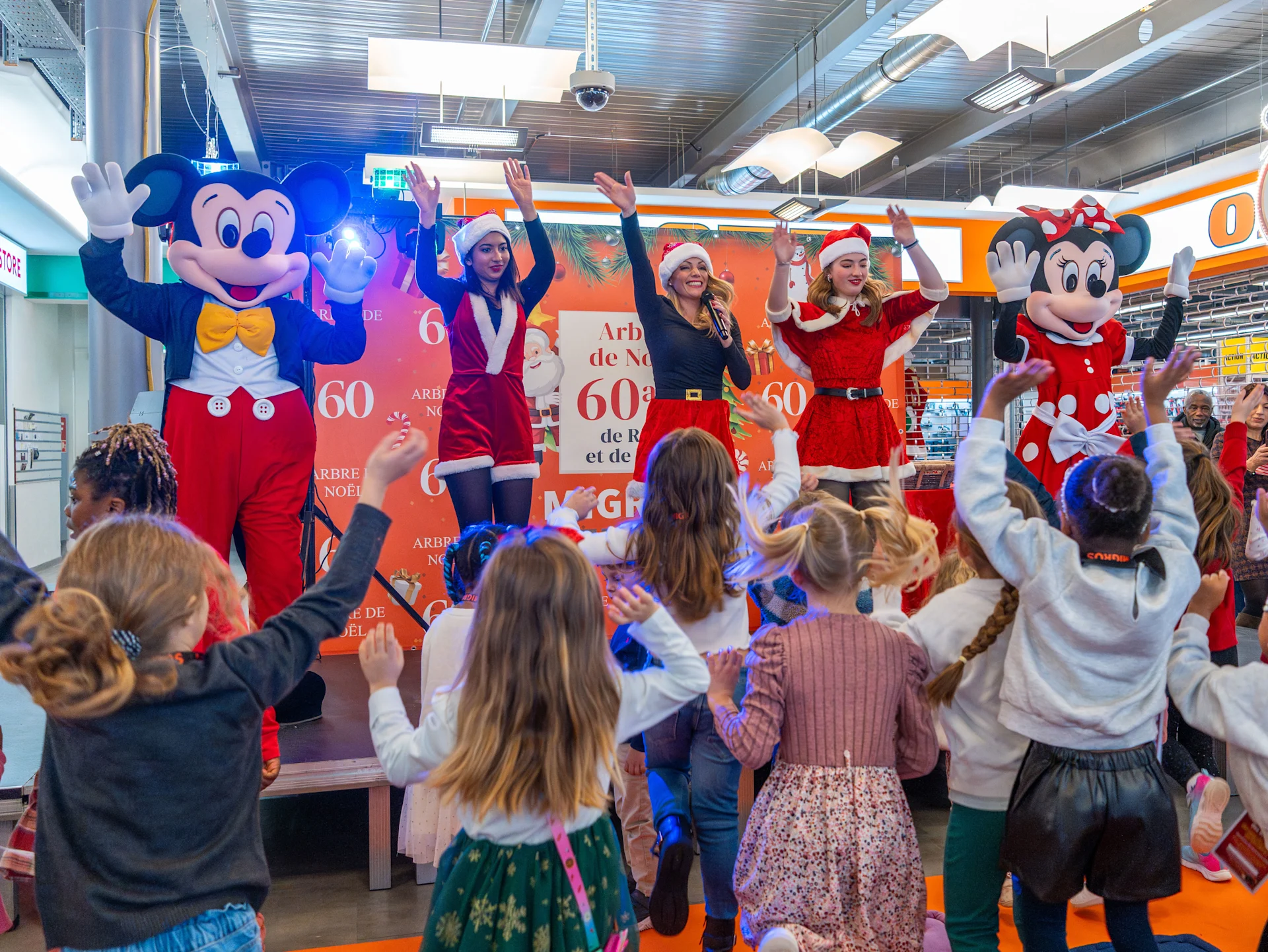 Mickey et 3 femmes avec des vêtements de Noël qui dansent devant une foule d'enfants.