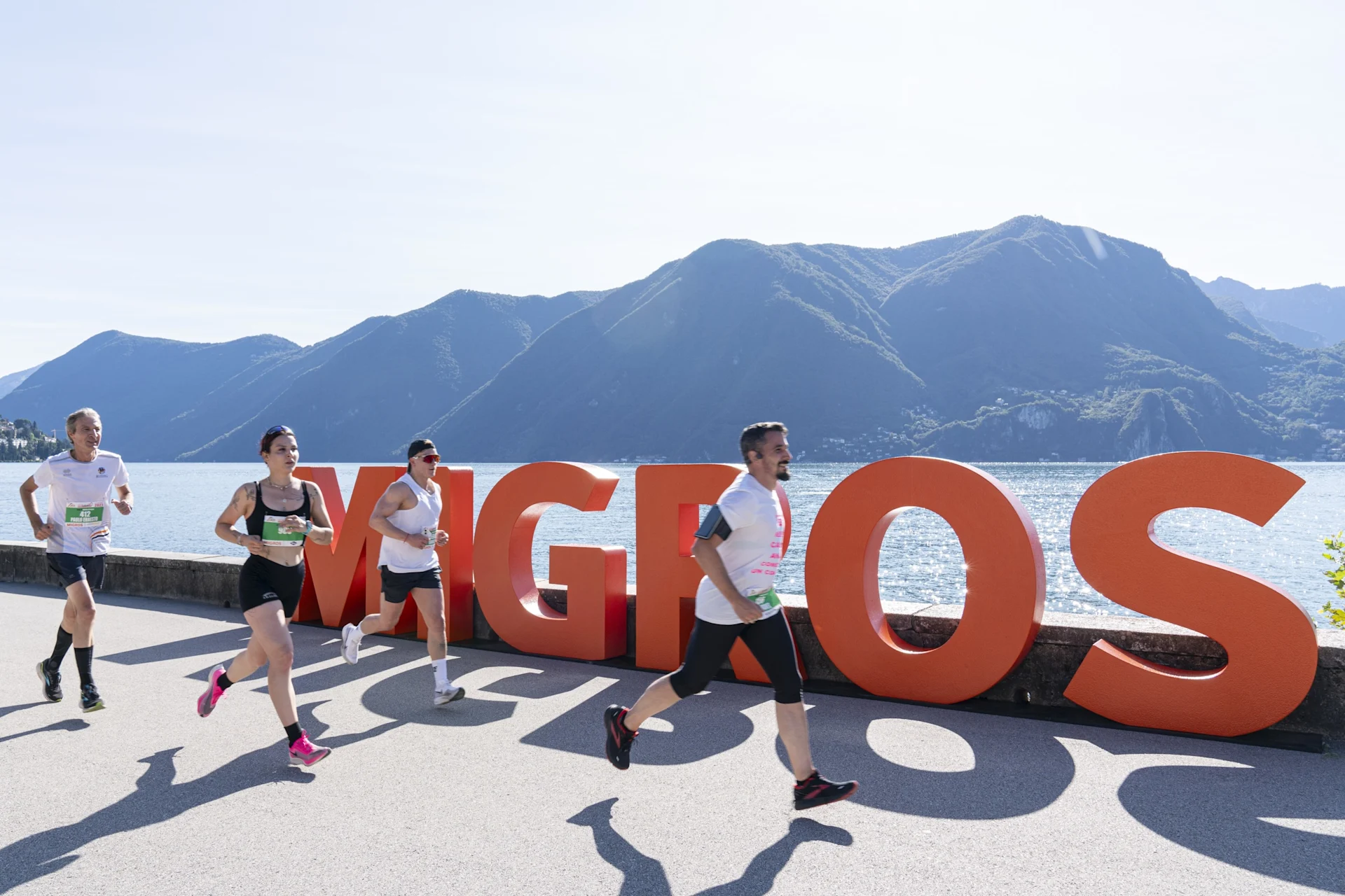 Verschiedene Personen joggen an einem See entlang, im Hintergrund der Migros-Schriftzug