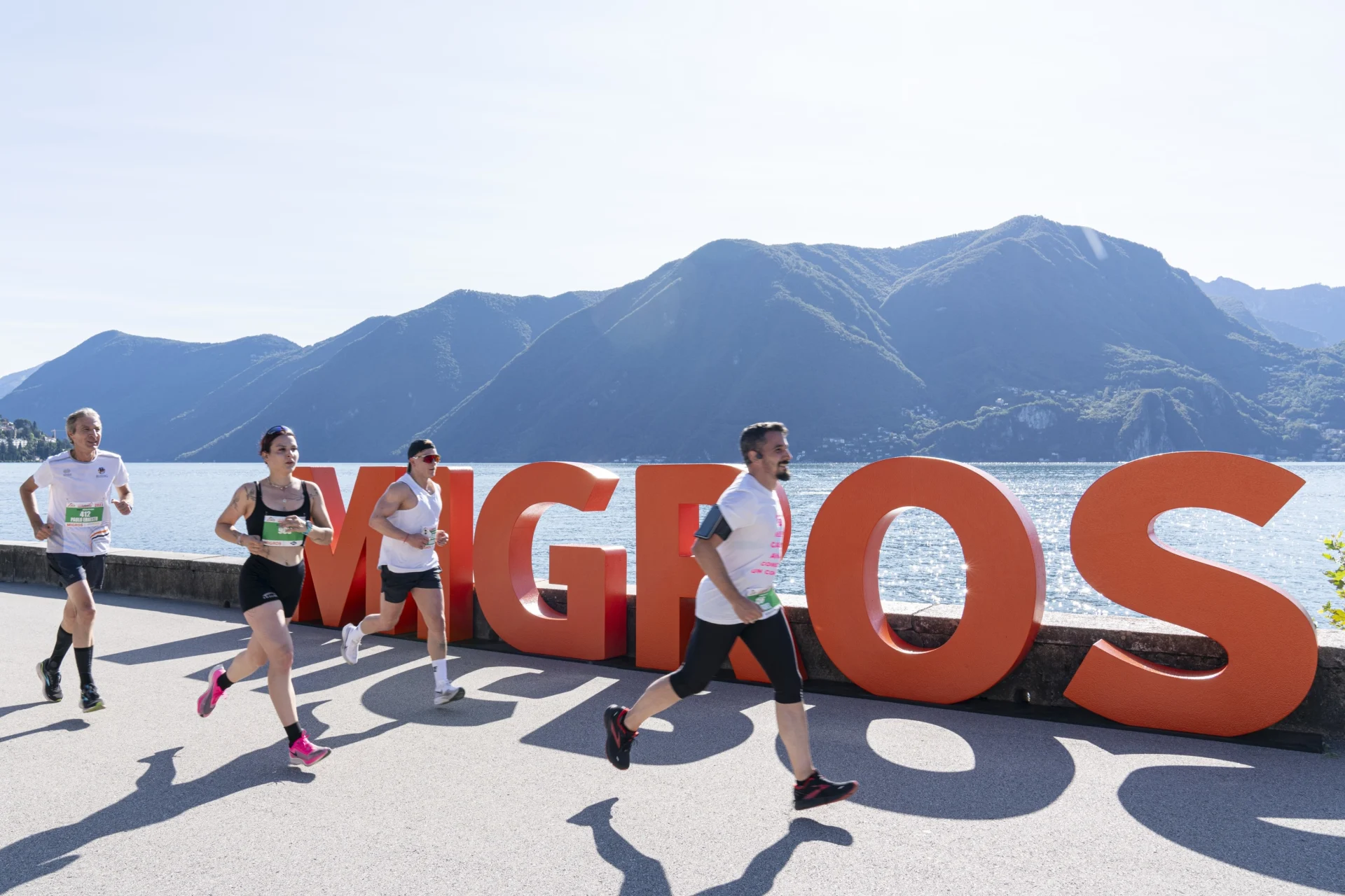 Various people are jogging along a lake, with the Migros logo in the background