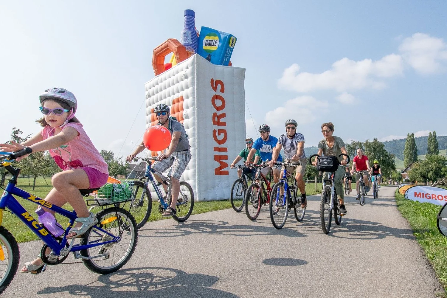 Several people are riding their bikes in front of a large inflatable Migros shopping bag.