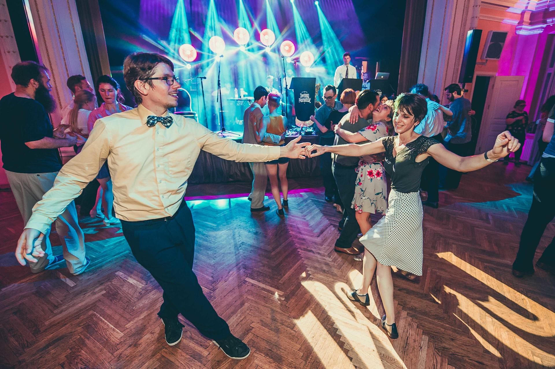 A couple is dancing the Lindy Hop in a ballroom in the foreground, a band is playing live music in the background.