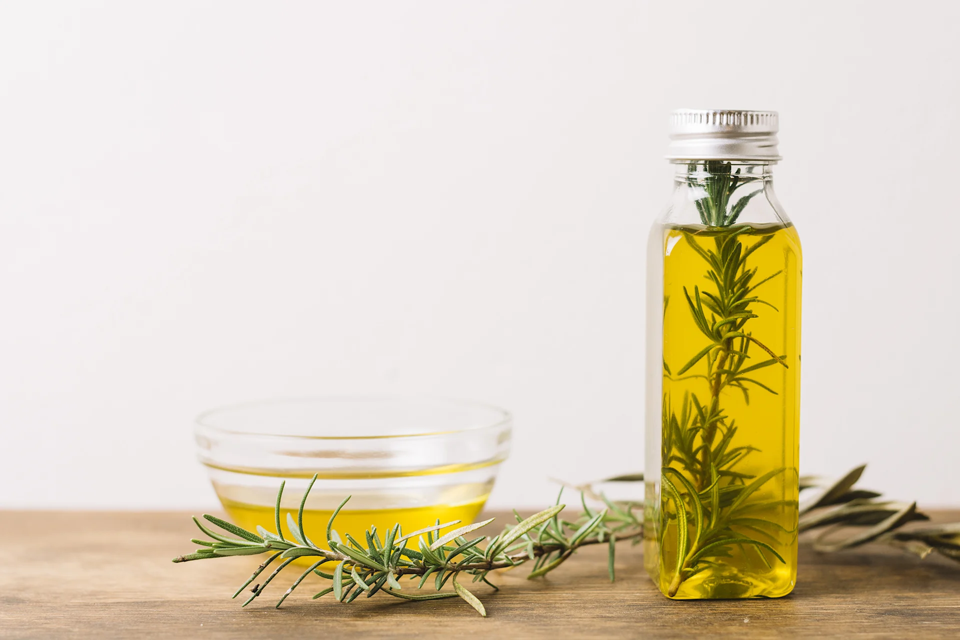 A small bowl of oil stands on the left-hand side on a wooden table. To the right, there is a small oil-filled bottle with a sprig of rosemary in it.