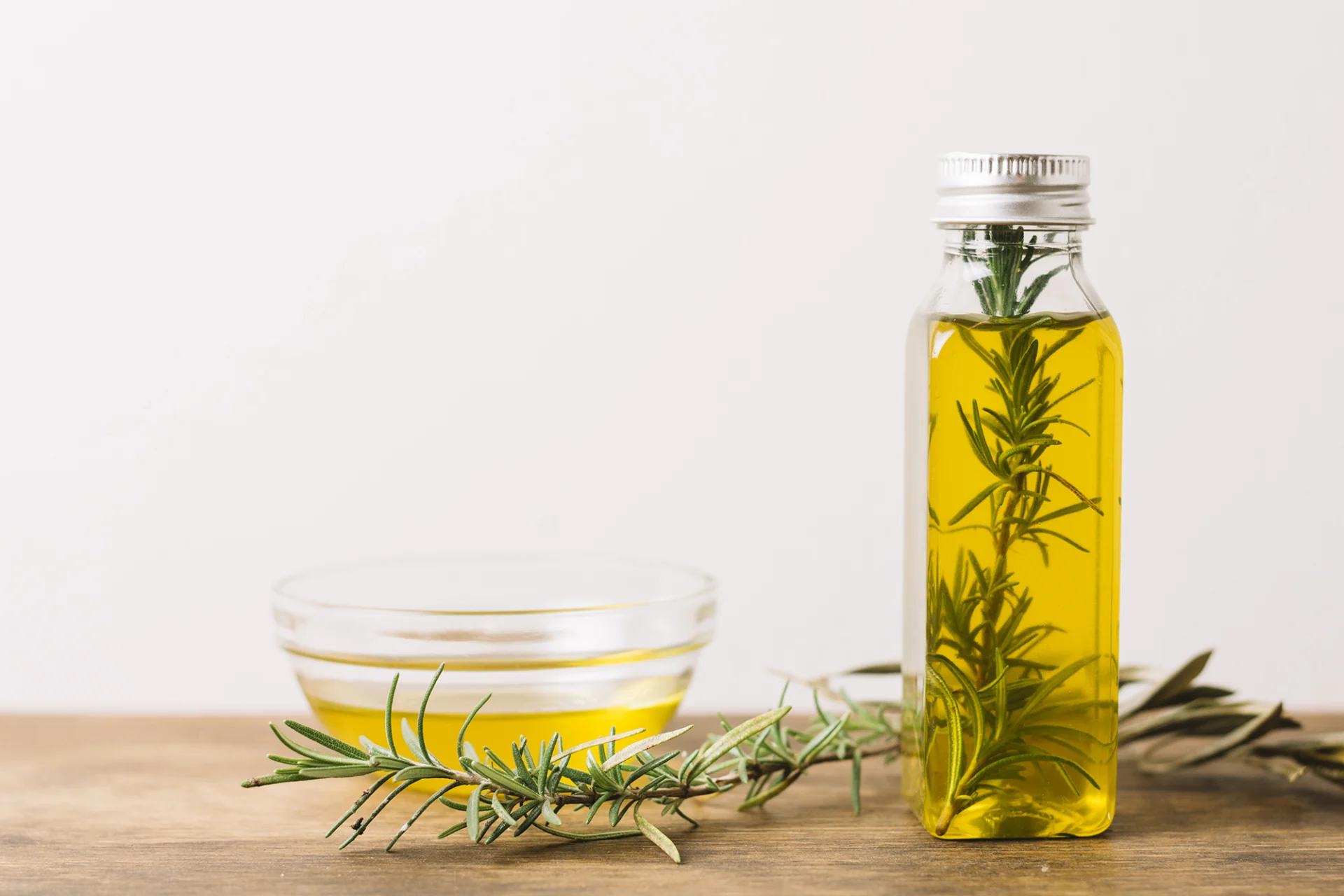 A small bowl of oil stands on the left-hand side on a wooden table. To the right, there is a small oil-filled bottle with a sprig of rosemary in it. 