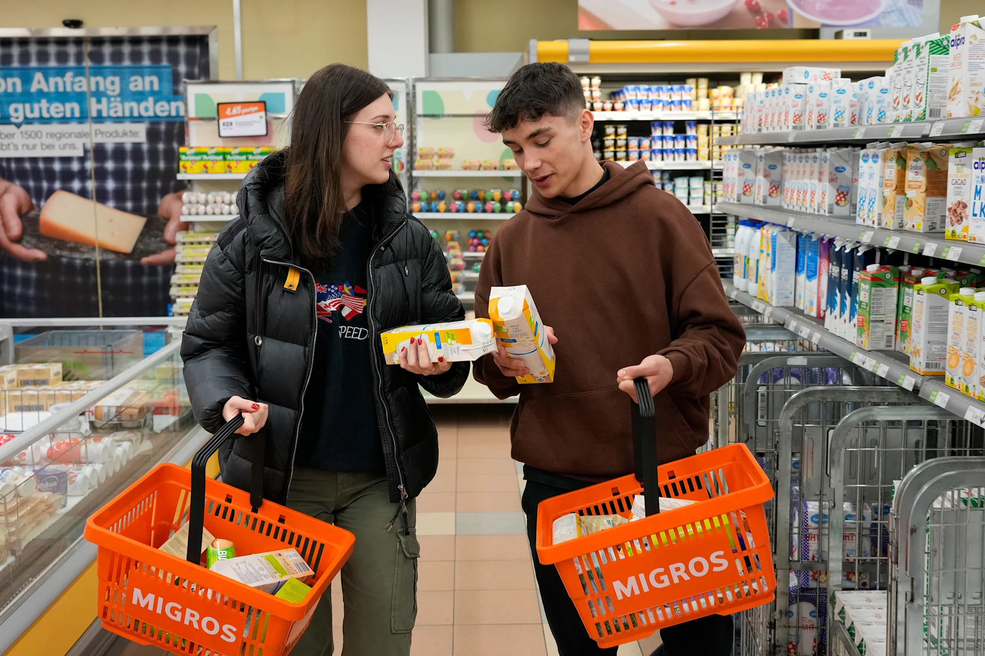 Une adolescente et un adolescent se tiennent entre des étagères Migros et tiennent des produits dans leurs mains