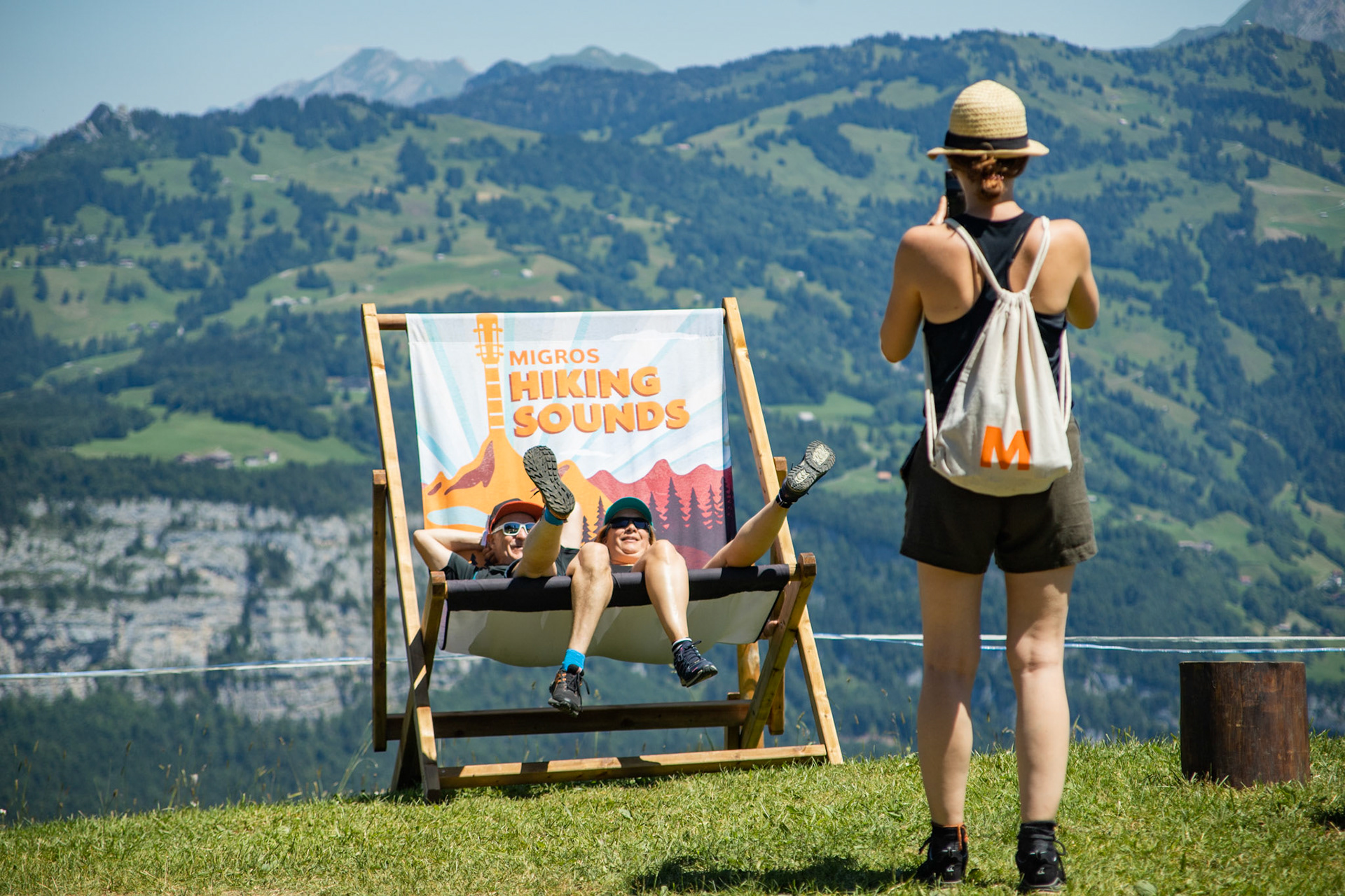 Une femme photographie deux personnes dans une chaise pliante démesurément grande devant un panorama de montagne.