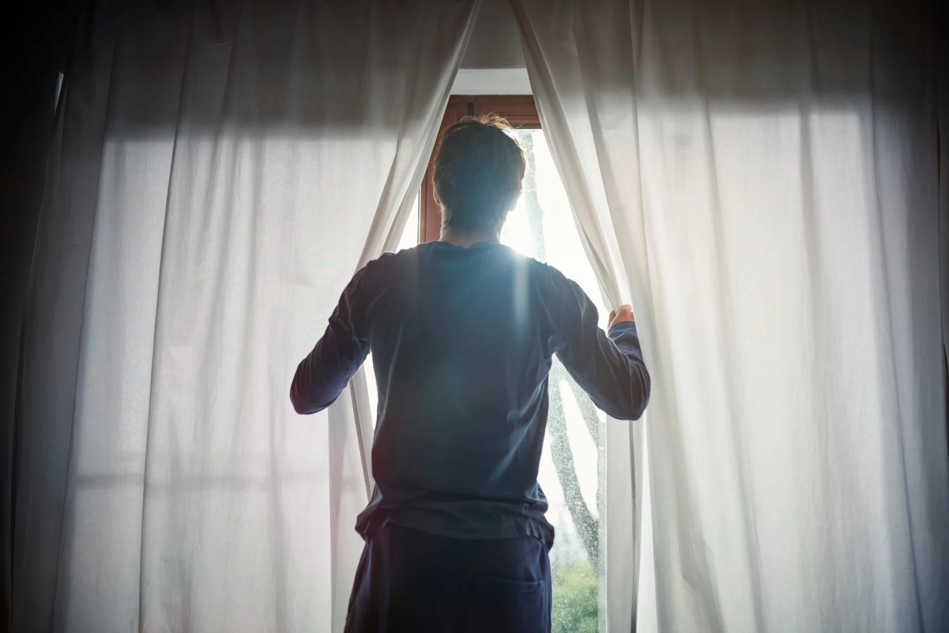 Ein Mann schiebt den Fenstervorhang zur Seite, um das Fenster zu öffnen.