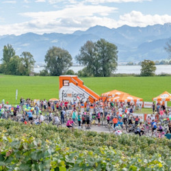 Famigros Familienevent im Grünen: Viele Teilnehmende mit Velos vor einem orangen Migros-Zelt und einer grossen aufblasbaren Rutschbahn, im Hintergrund Wiese, See und Berge.