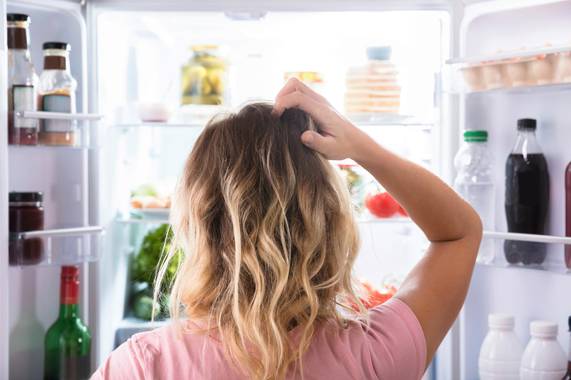 A woman stands in front of an open fridge, scratching her head