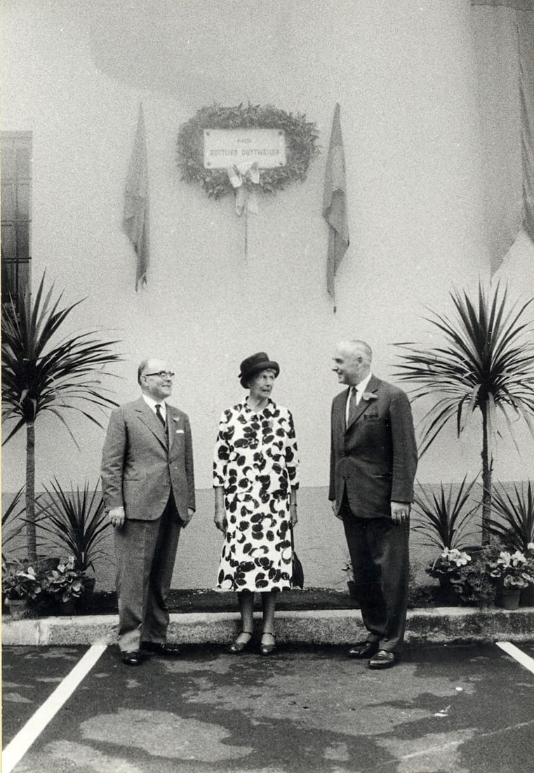 Adele Duttweiler und die Herren Guglielmetti und Hochstrasser vor dem Schild der Piazza Gottlieb Duttweiler (1963).  [MGB_Dok_Fo_139405]