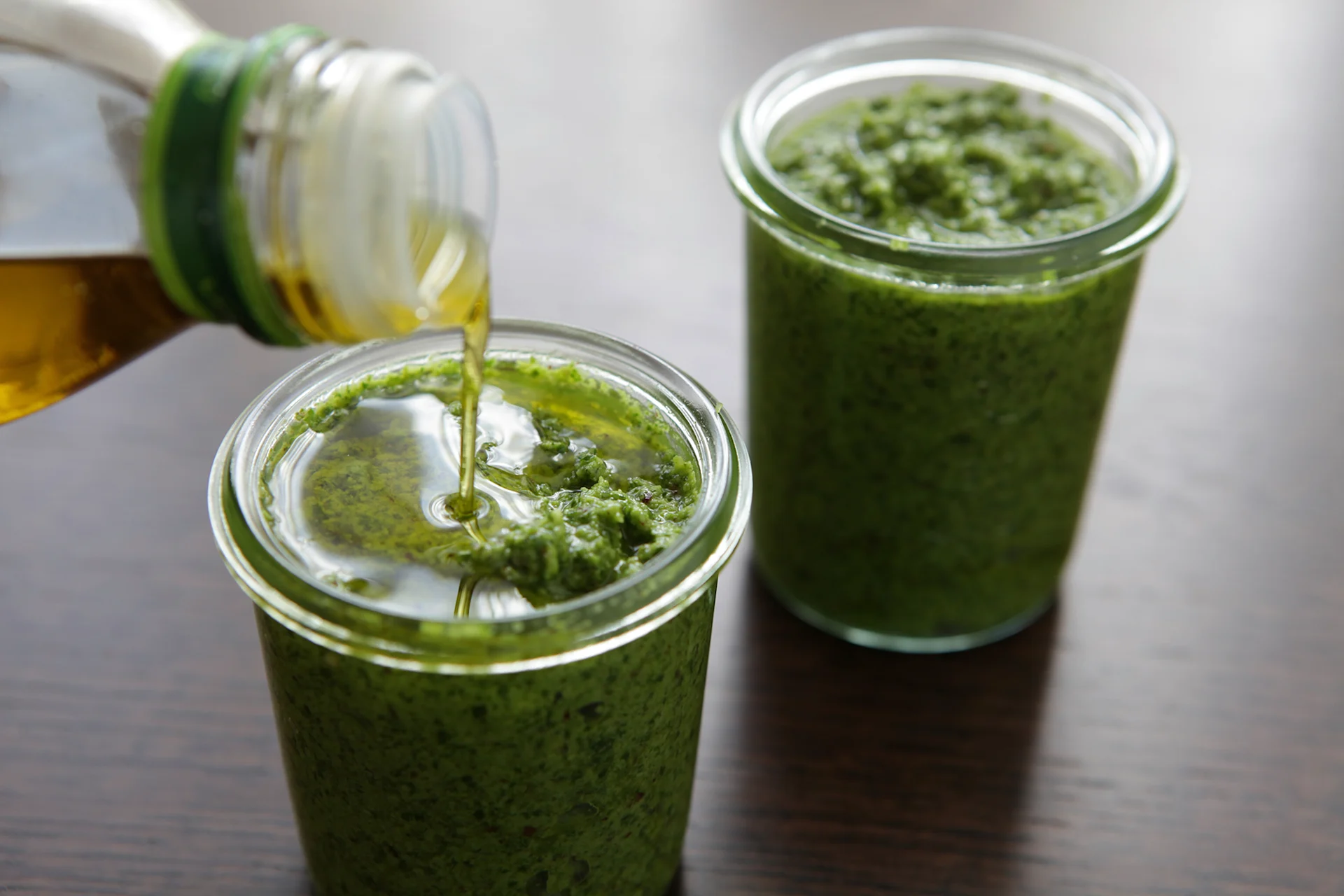 Two small jars filled with rocket pesto. Some oil is being poured into the jar on the left.