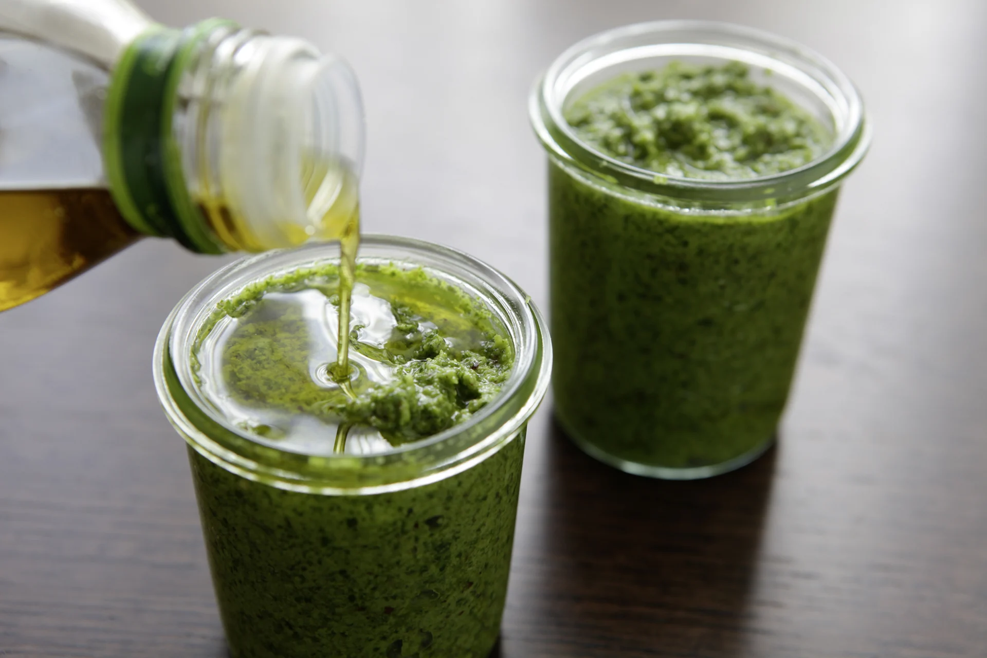 Two small jars filled with rocket pesto. Some oil is being poured into the jar on the left.