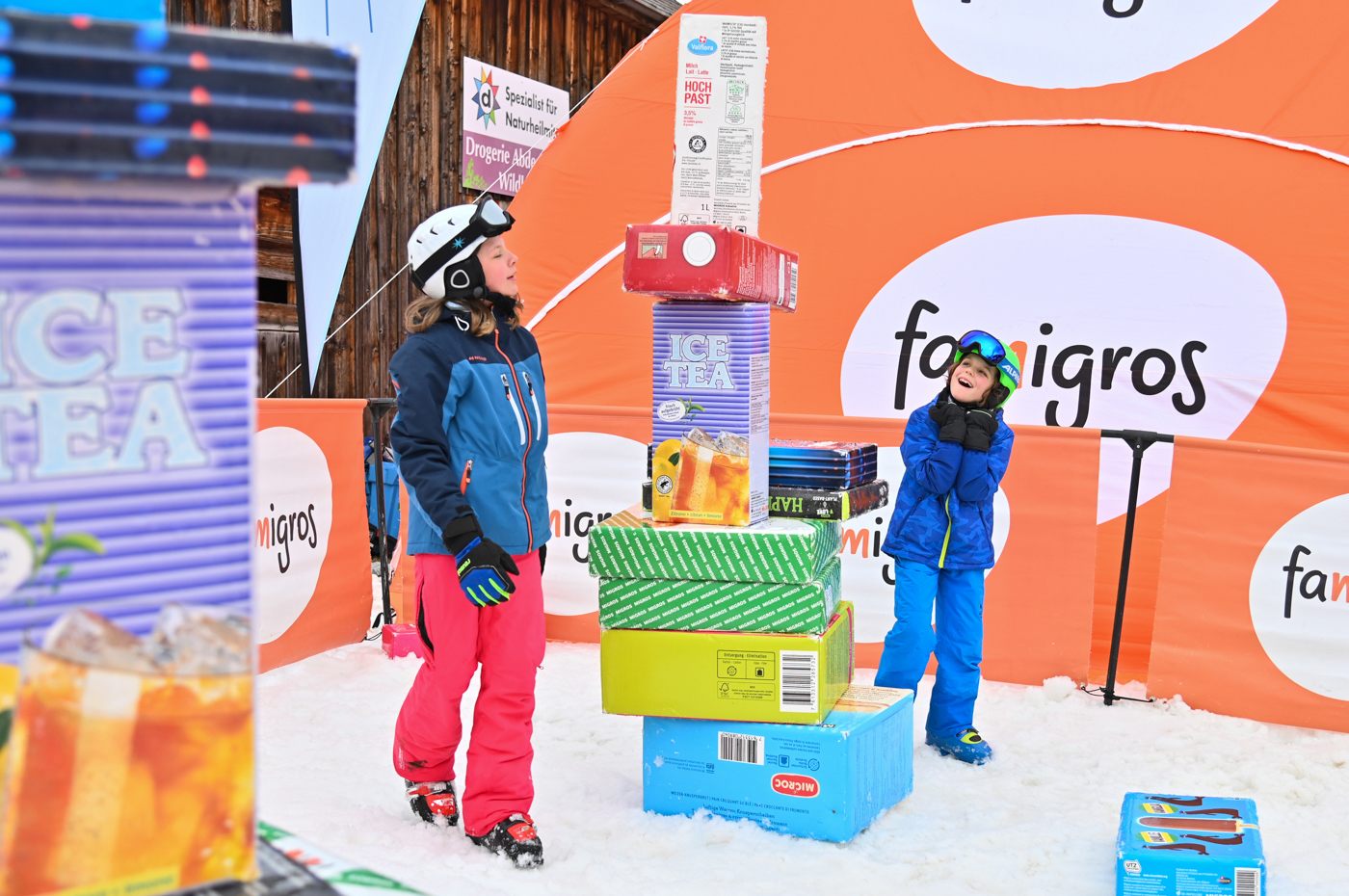 Due ragazze impilano sulla neve confezioni Migros giganti, come l'Ice Tea.