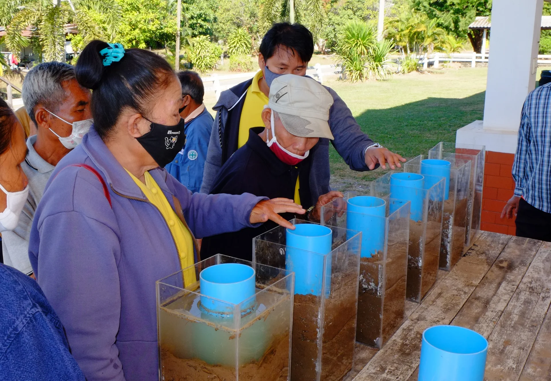 Thailändische Bäuer*innen sehen sich durchsichtige, mit Wasser und Erde gefüllte Behälter an.