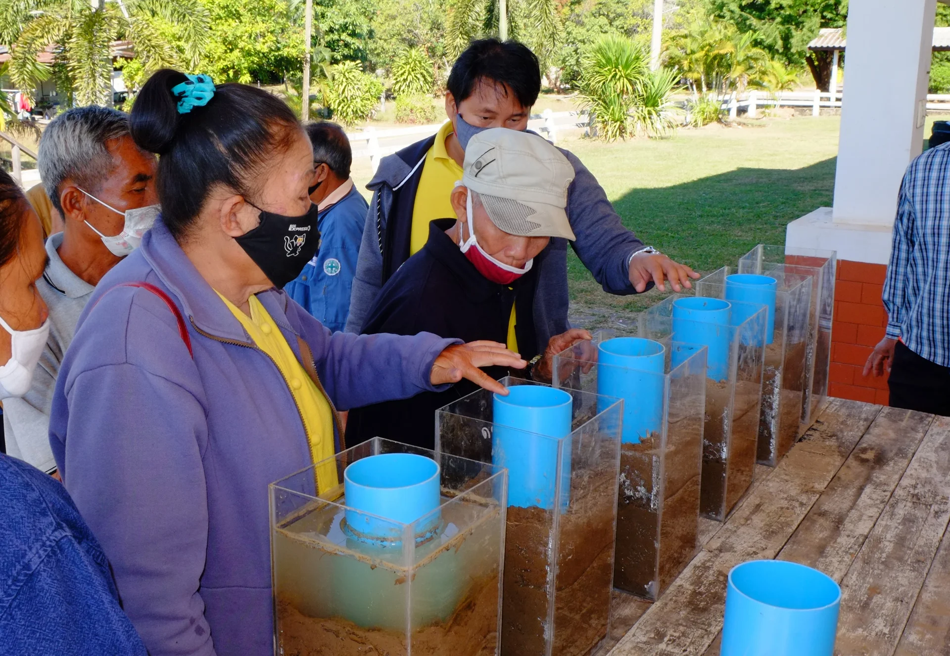 Des paysannes et paysans thaïlandais observent des récipients transparents remplis d’eau et de terre.