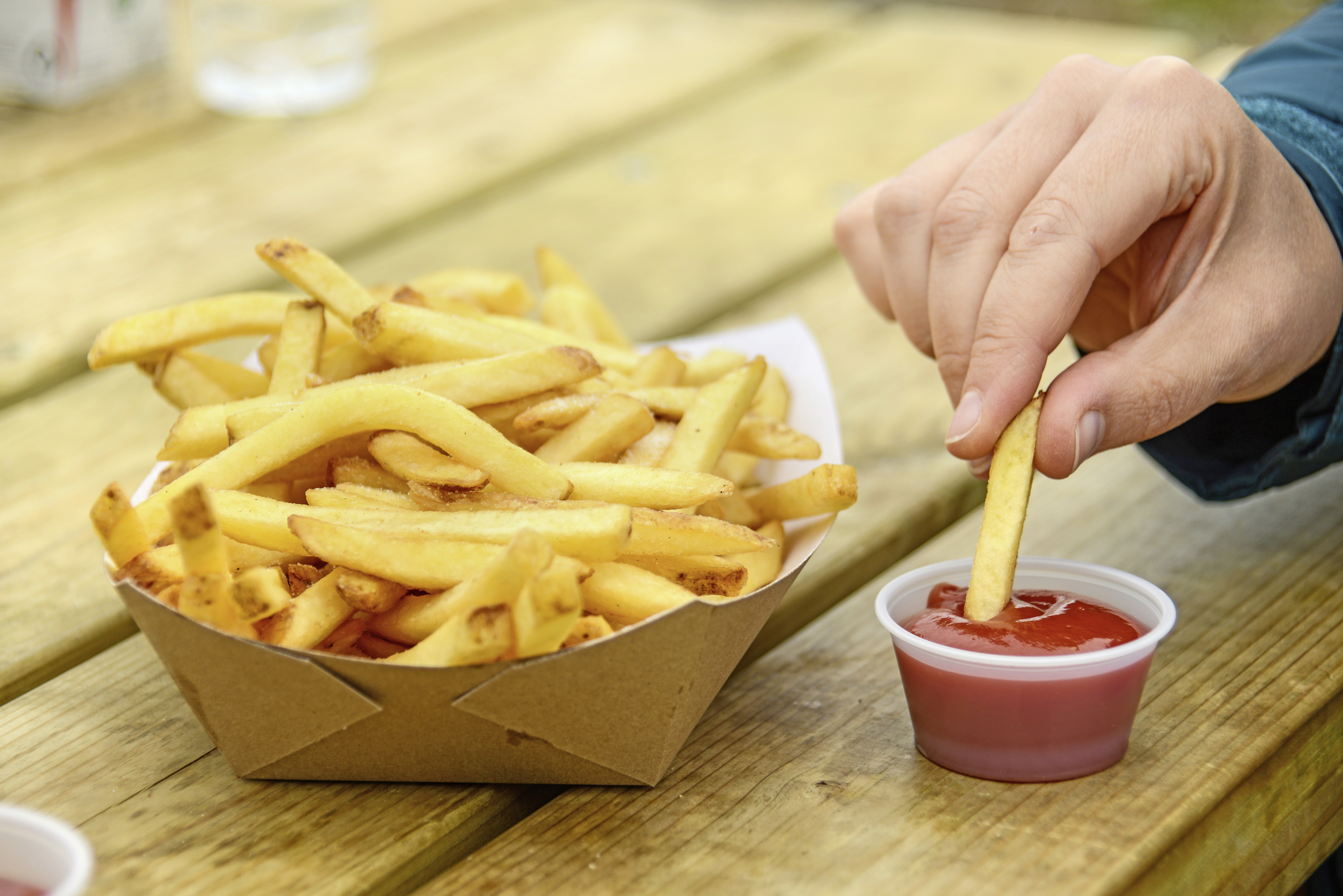 A serving of chips on a table, a hand takes one and dips it in ketchup.