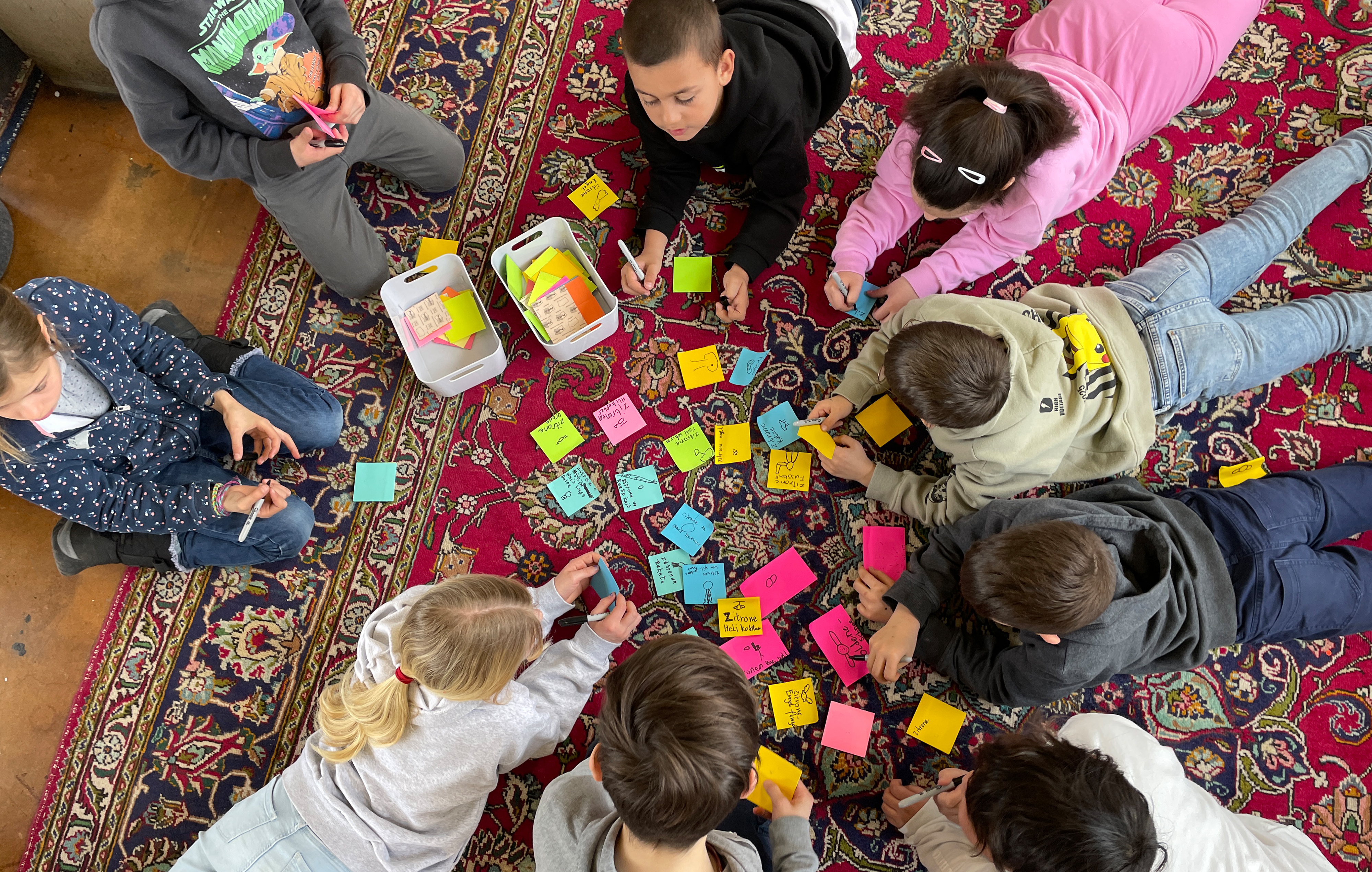 Mehrere Kinder liegen im Kreis auf dem Boden. In ihrer Mitte sind viele farbige Post-its ausgebreitet.