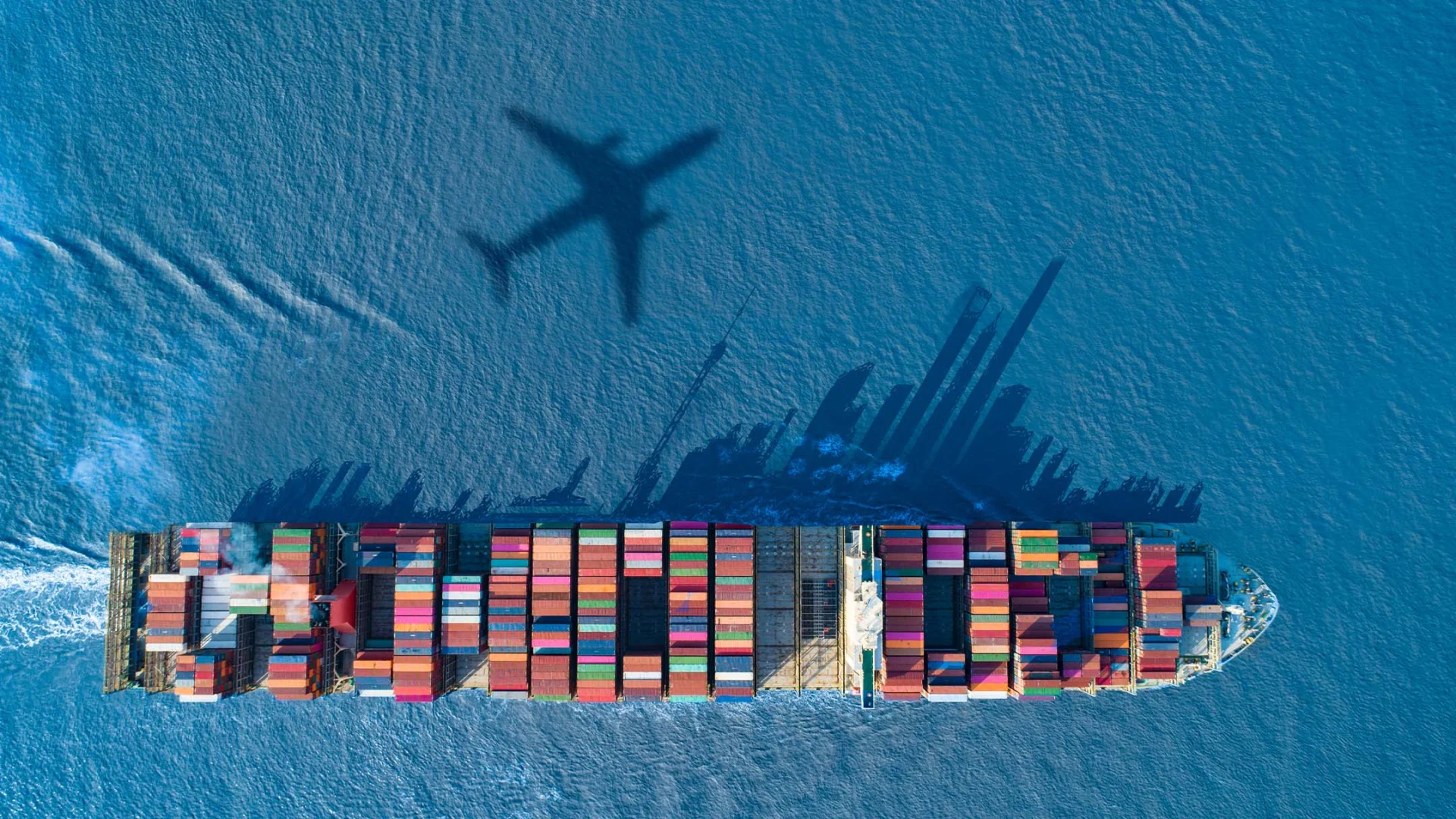Ein Frachtschiff im Meer von oben, darüber der Schatten eines Flugzeugs