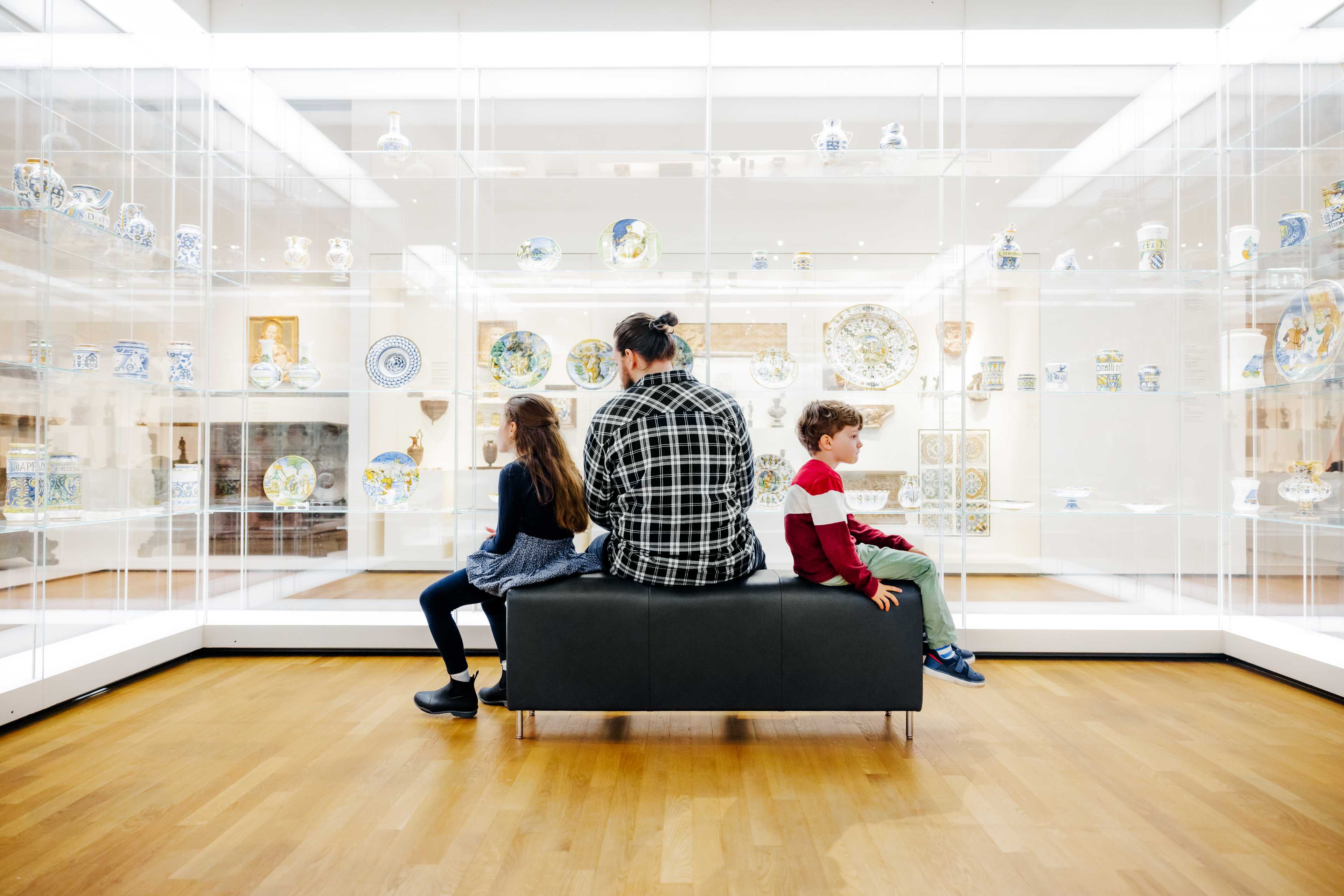 Un père et ses deux enfants sont assis sur un tabouret dans une salle d’œuvres d’art.